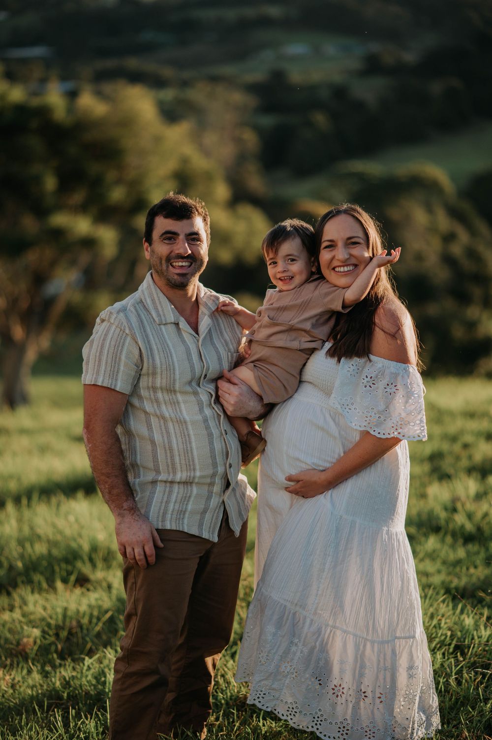 A family stands together in a sunlit field at golden hour, with the expecting mother wearing a white eyelet dress.