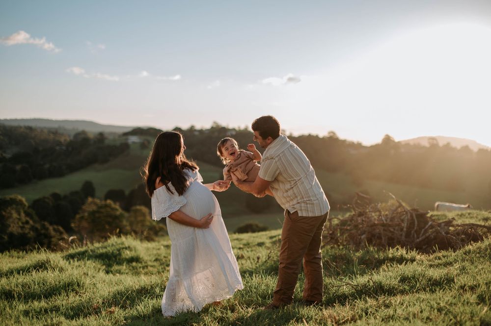 Two people share a joyful moment with a small child on a grassy hilltop during golden hour sunset.