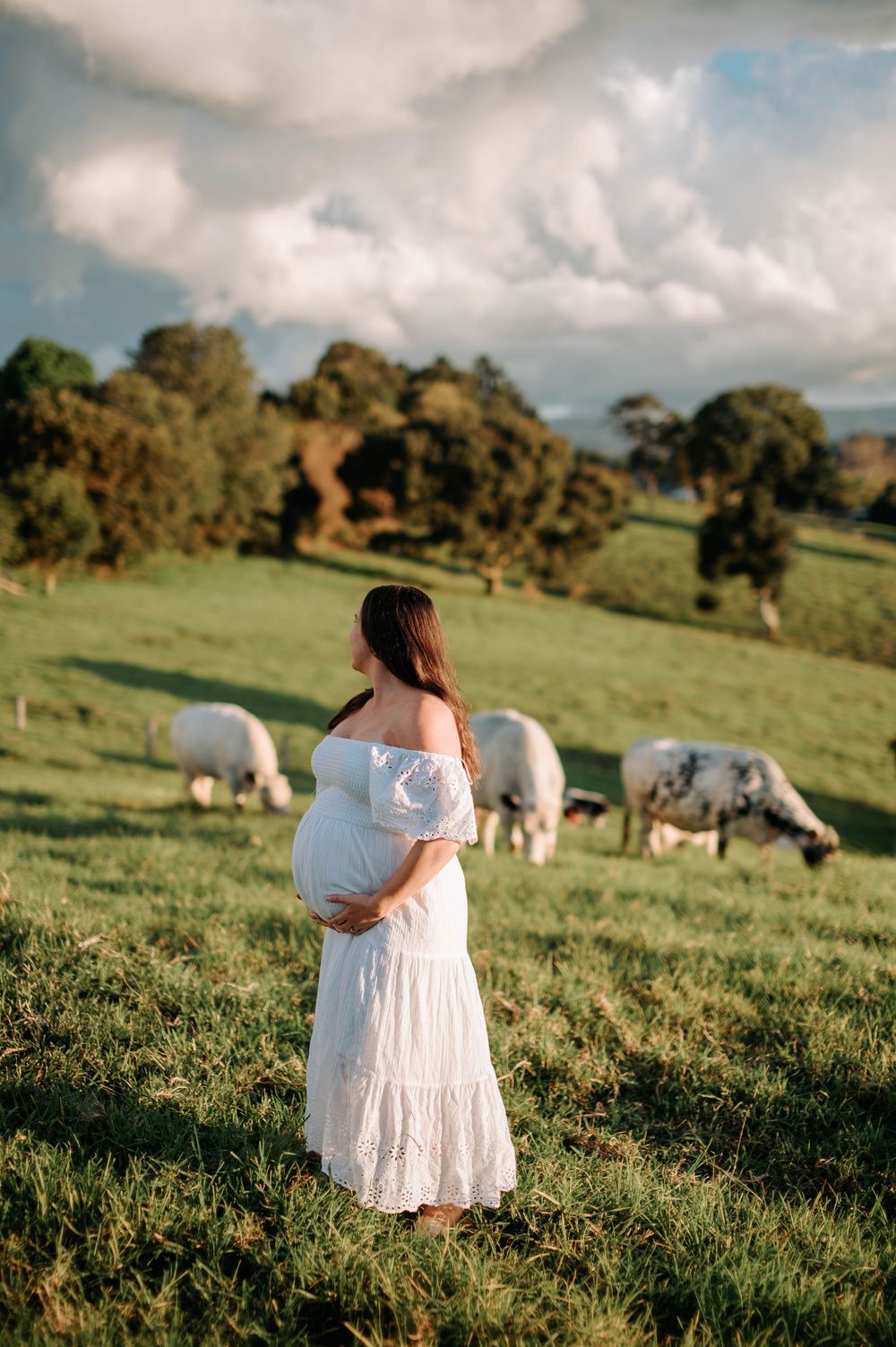 A pregnant woman in a white dress stands in a field with grazing sheep on a cloudy day.