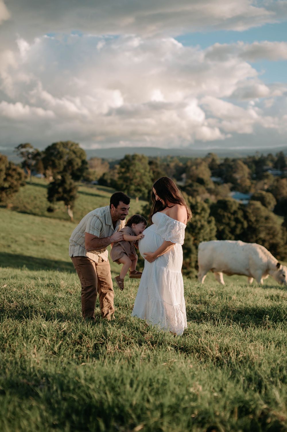 A family shares a tender moment in a pastoral field with rolling hills and a white cow grazing in the background.