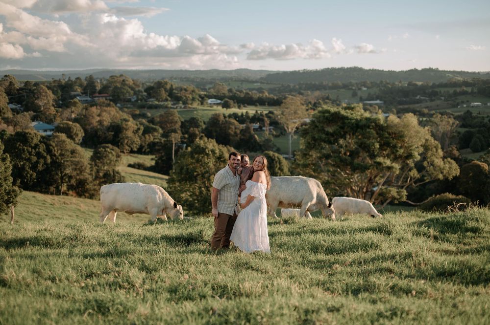 A couple shares an intimate moment in a green pasture with white horses grazing in the countryside background.