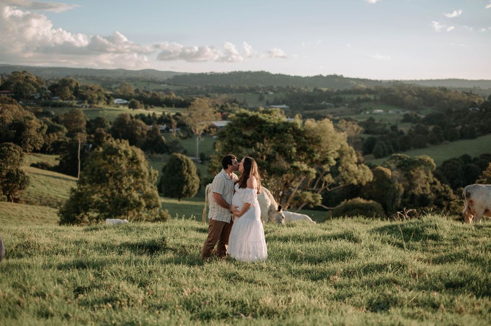 A couple stands together on a grassy hillside overlooking a scenic countryside landscape at sunset.