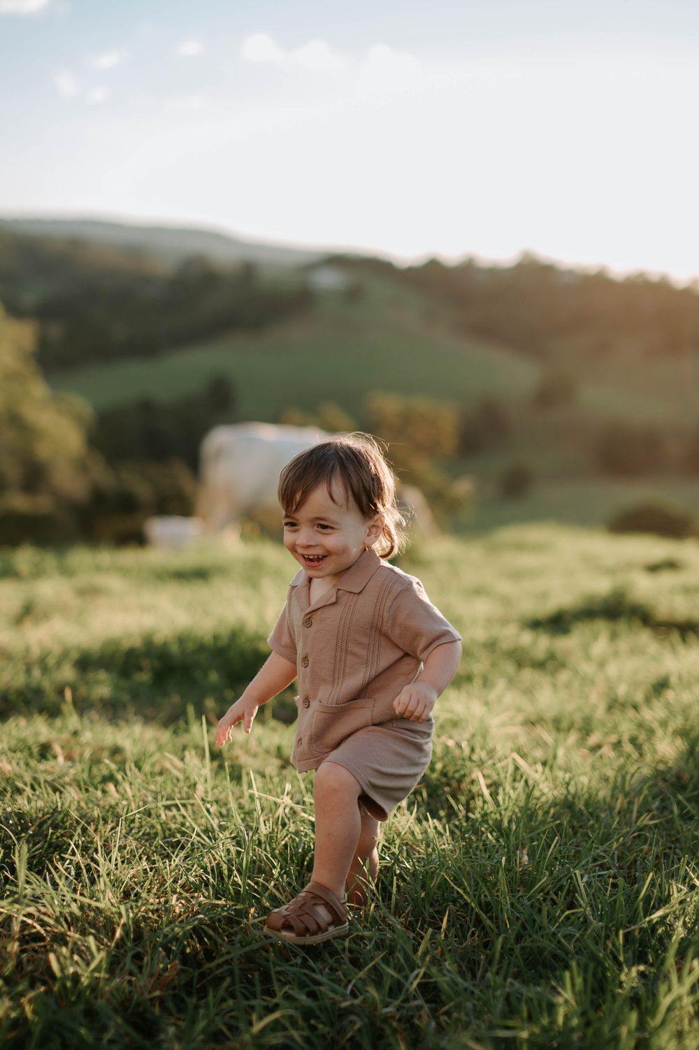 Small toddler wearing tan linen outfit plays in sunny green hillside meadow at golden hour.