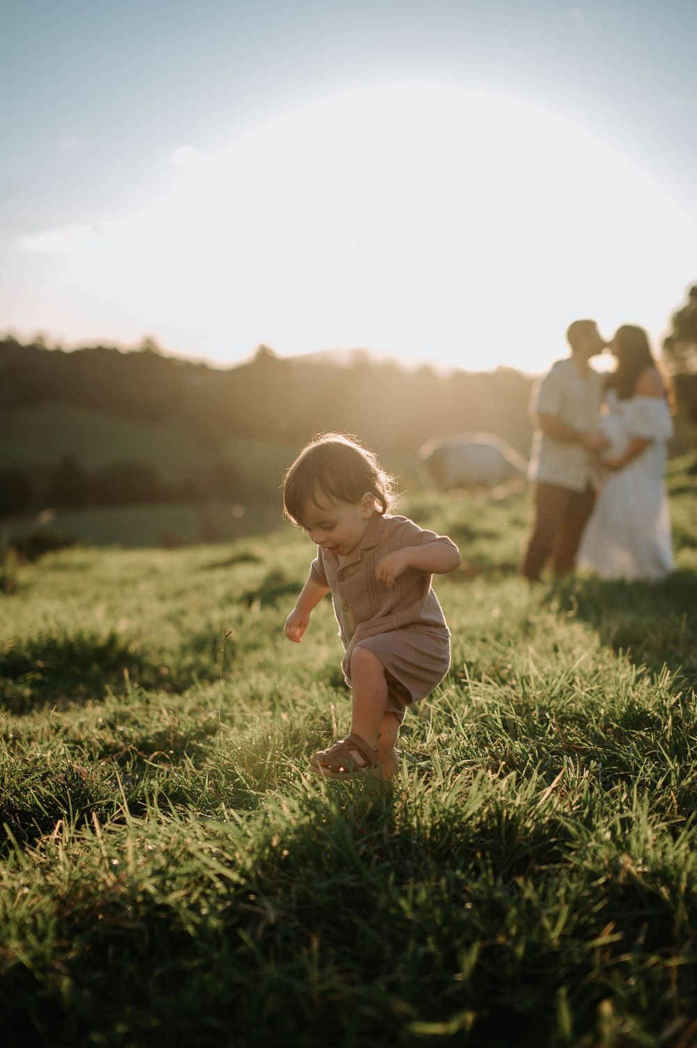 Small child in brown romper plays in a grassy field during golden hour sunset with blurred figures in background.