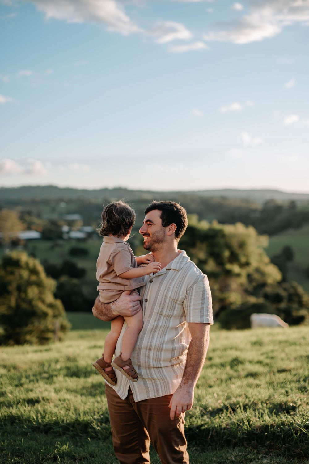 Parent and child share a joyful moment together on a grassy hillside against a blue sky and scenic landscape.