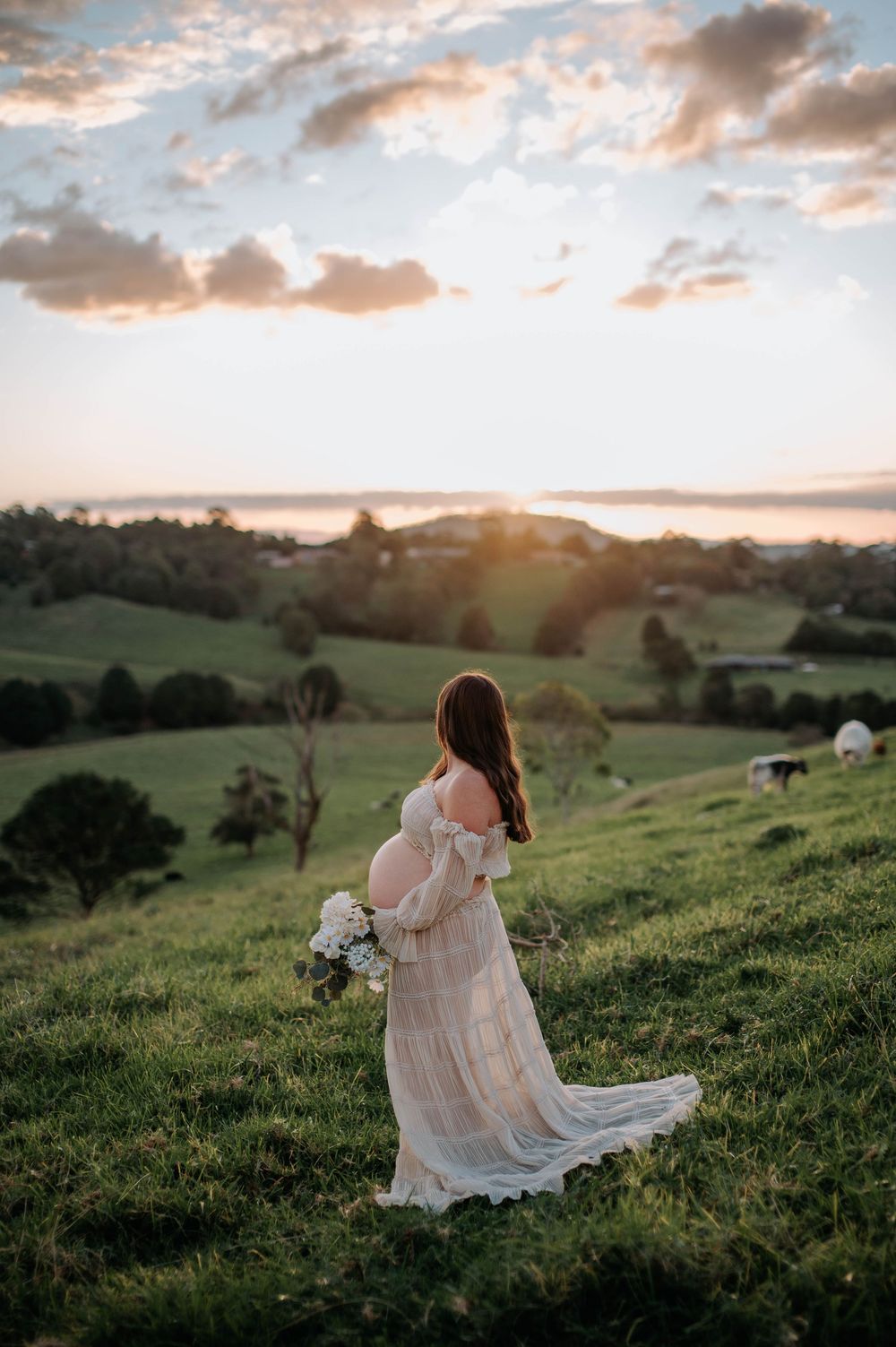 A graceful figure in a flowing white dress holds a bouquet while standing on a hilltop at sunset.