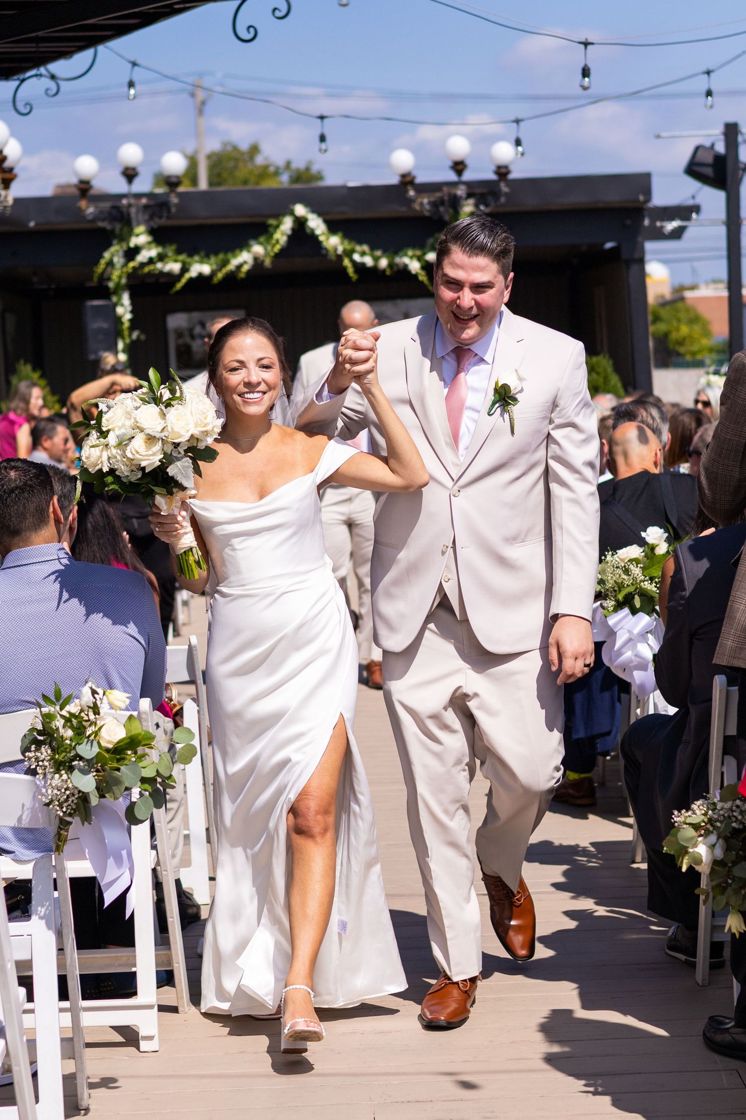 Happy bride and groom walk hand-in-hand down the aisle during their sunny outdoor wedding ceremony.