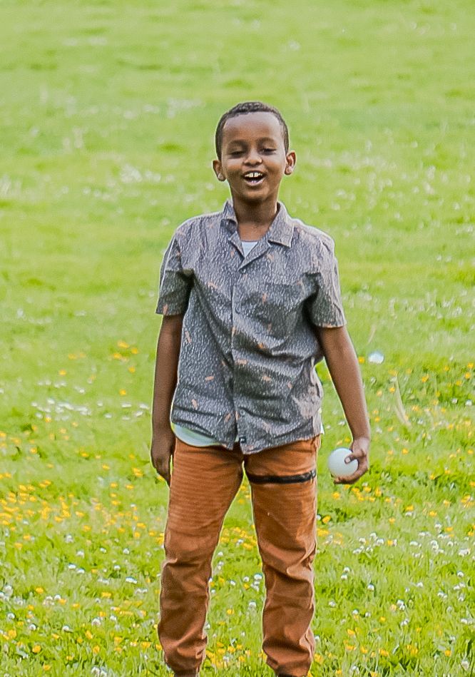 Young child in a gray button-up shirt and brown pants smiling joyfully while standing on grassy field in summer.