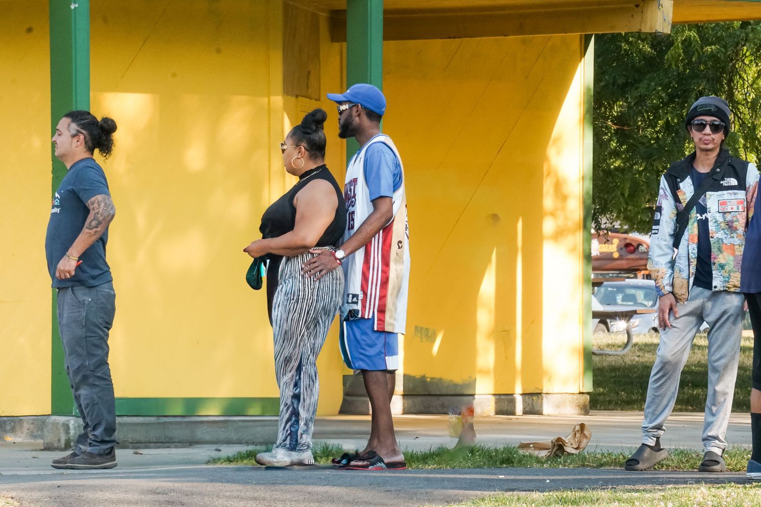 People waiting in line outside a yellow building with teal trim on a sunny day.