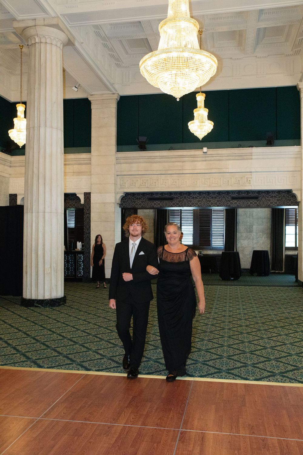 Couple in formal black attire walking across an elegant ballroom with crystal chandeliers and marble columns.
