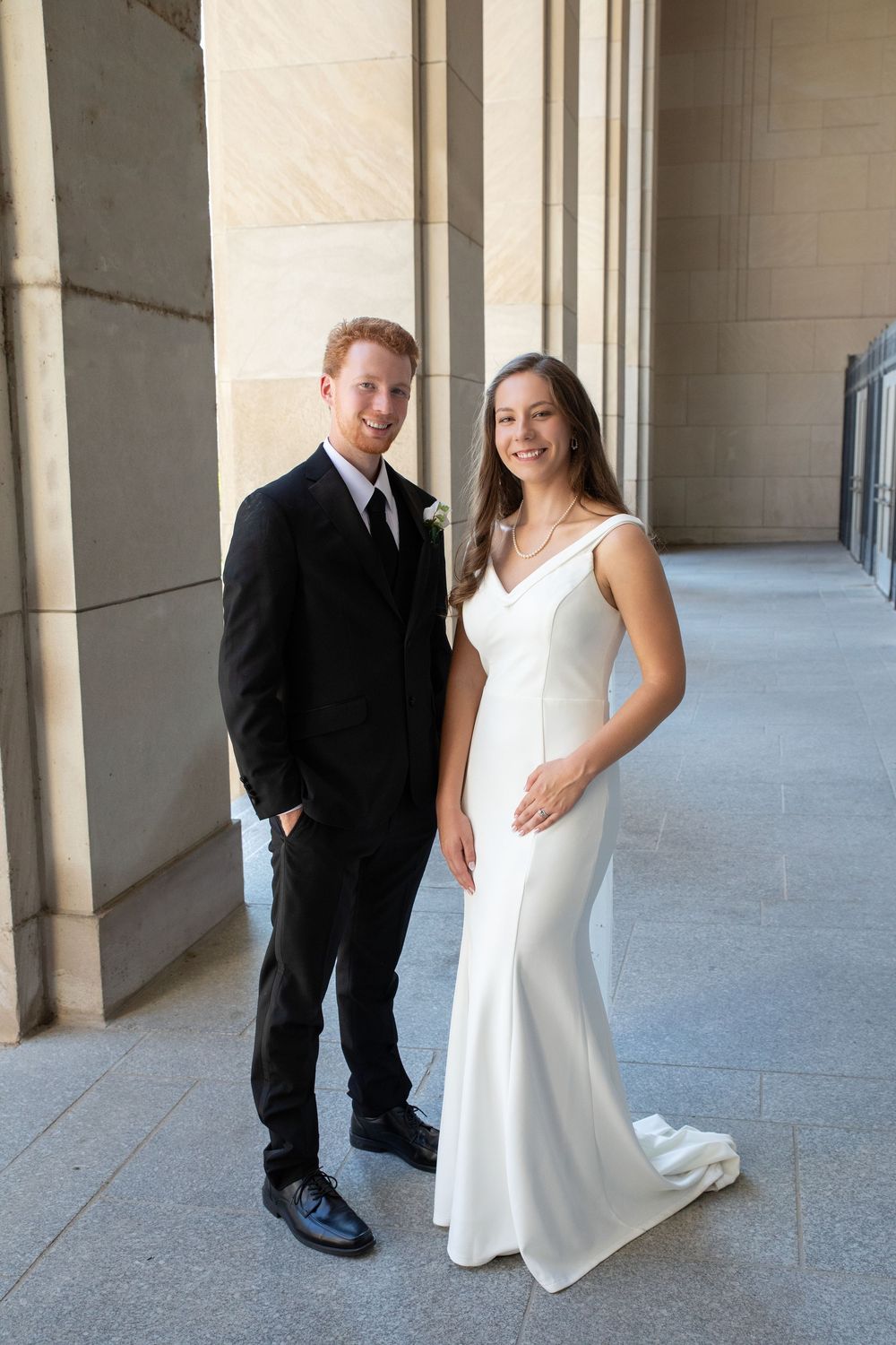 A couple poses in formal attire along a columned hallway with white stone architecture.