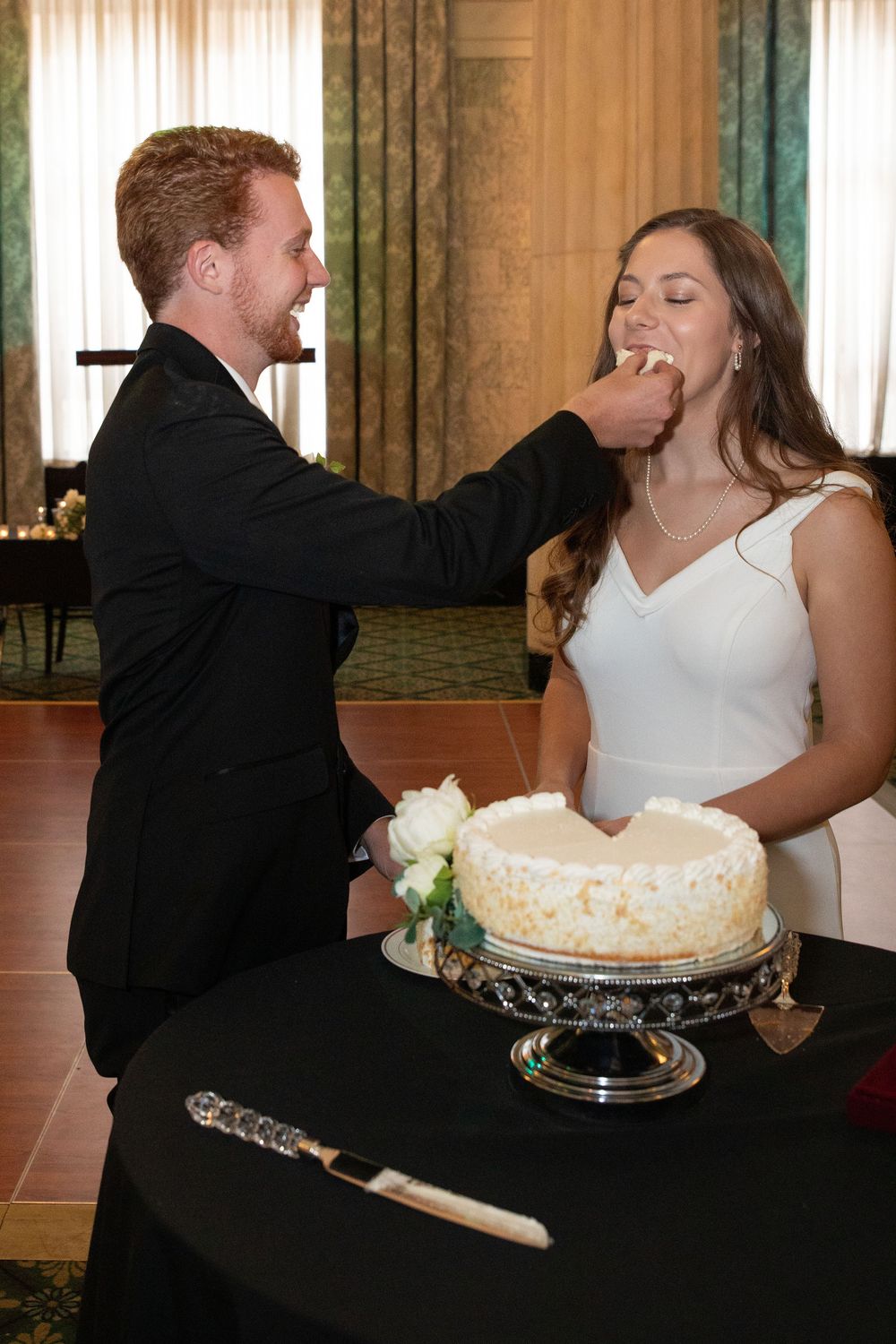 Newlyweds playfully share wedding cake at their reception with decorative serving pieces.