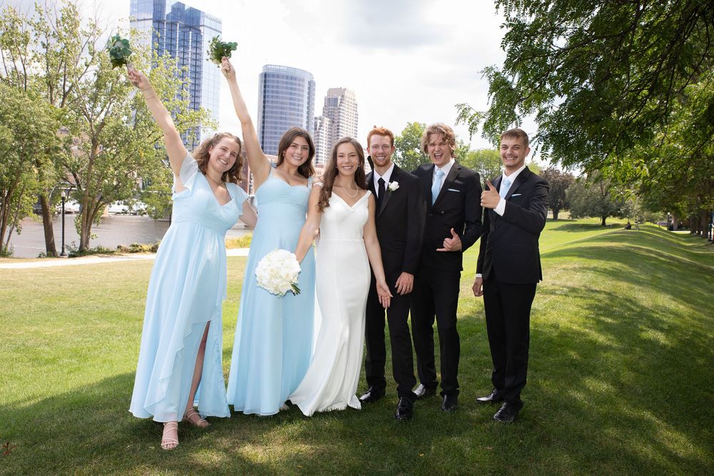 Wedding party in light blue bridesmaid dresses and black suits pose together on green lawn with city skyline behind them.