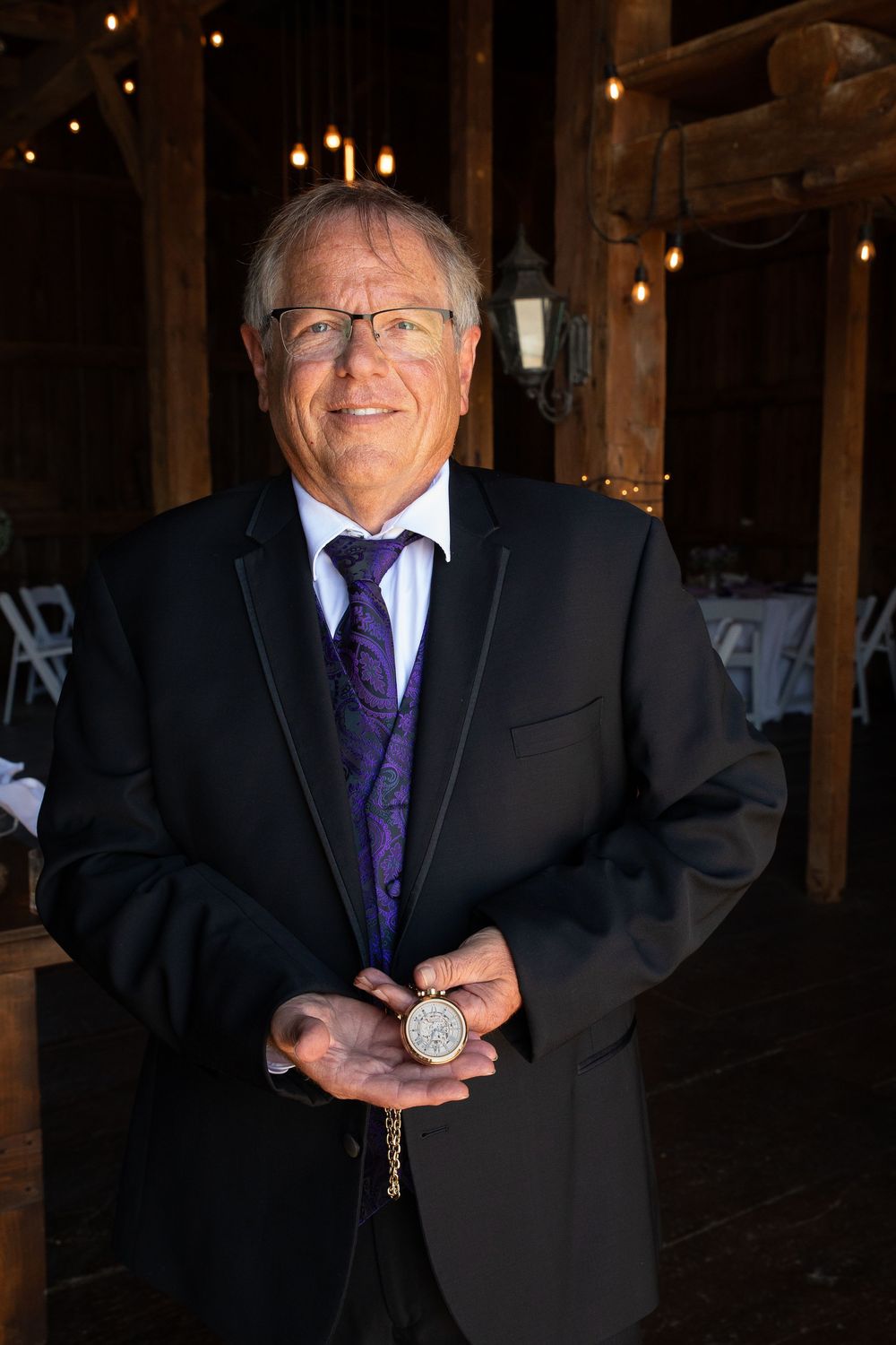 Smiling senior man in black suit and purple tie holding a gold pocket watch at an elegant barn venue.