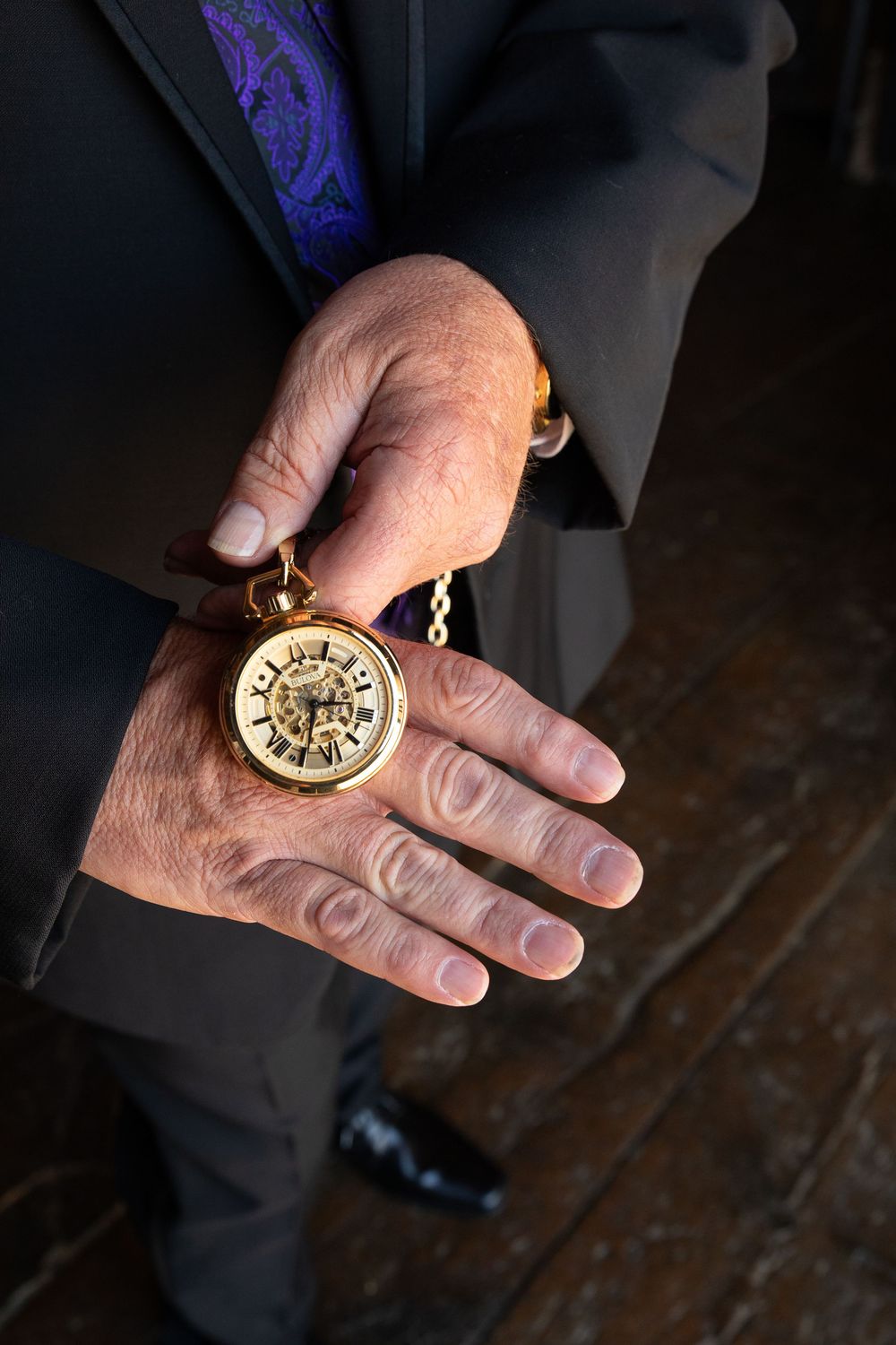 Elderly man in suit holding ornate gold pocket watch with chain in wrinkled hand.