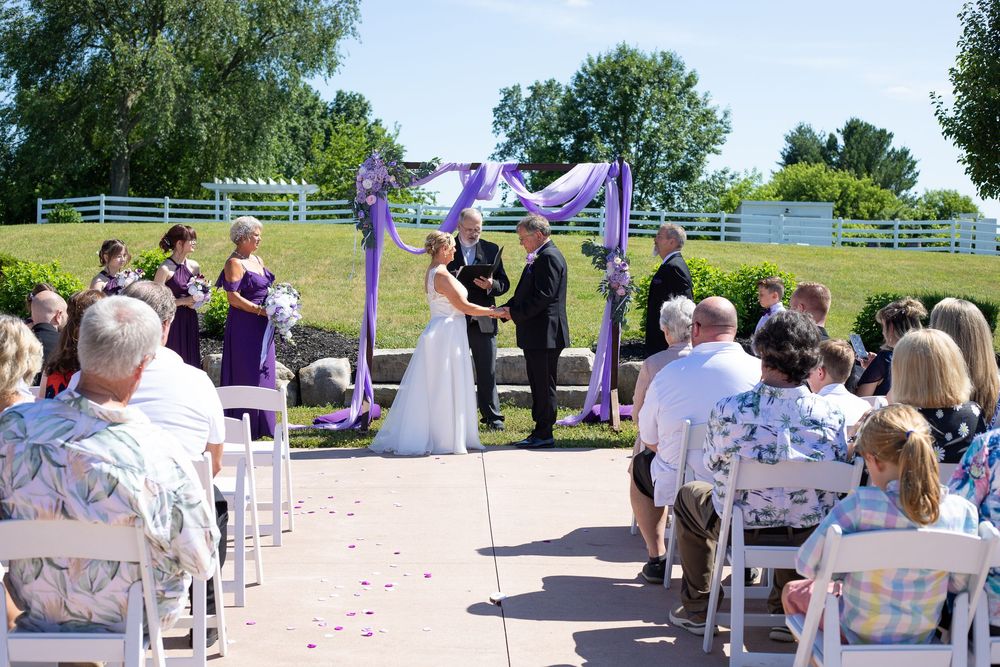 Outdoor wedding ceremony with bride and groom at purple-decorated arch, guests seated on white chairs.