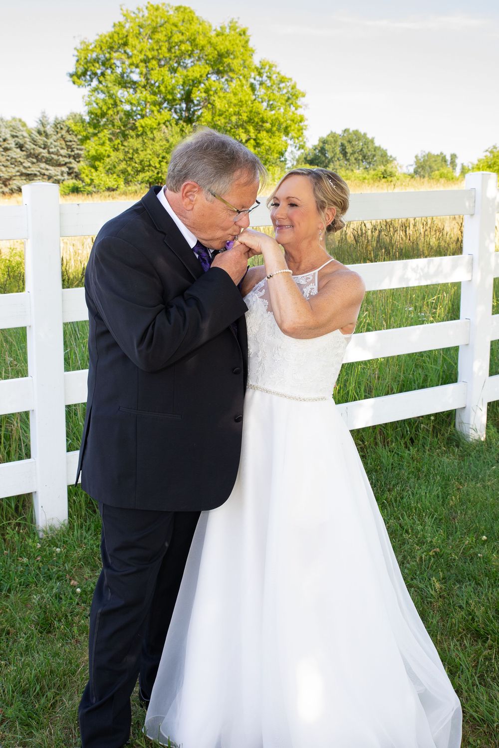 Older couple in wedding attire sharing a romantic hand kiss moment outdoors by a white fence.