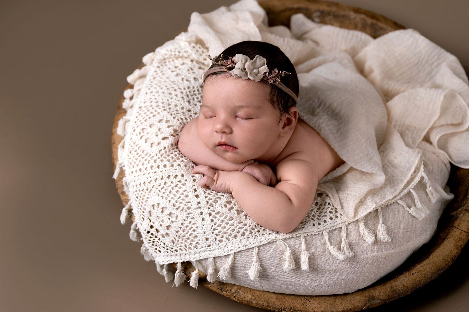 Sleeping newborn rests peacefully on white knitted blanket with delicate lace headband in brown toned portrait.