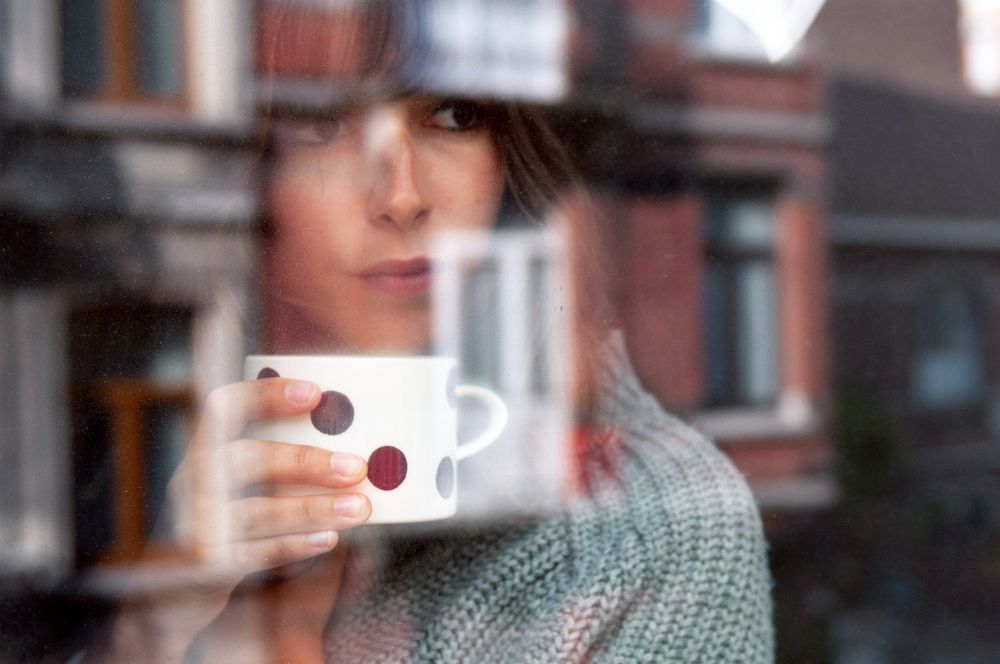 Person holding white coffee cup near a window with soft natural lighting.