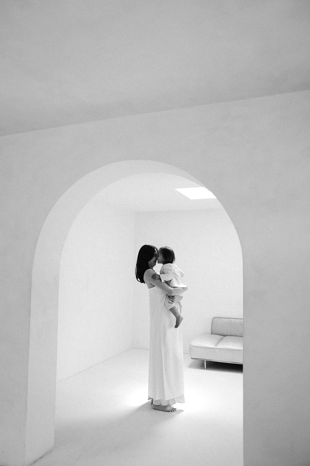Black and white photograph of a person in a white dress holding a small child under a minimalist archway interior.