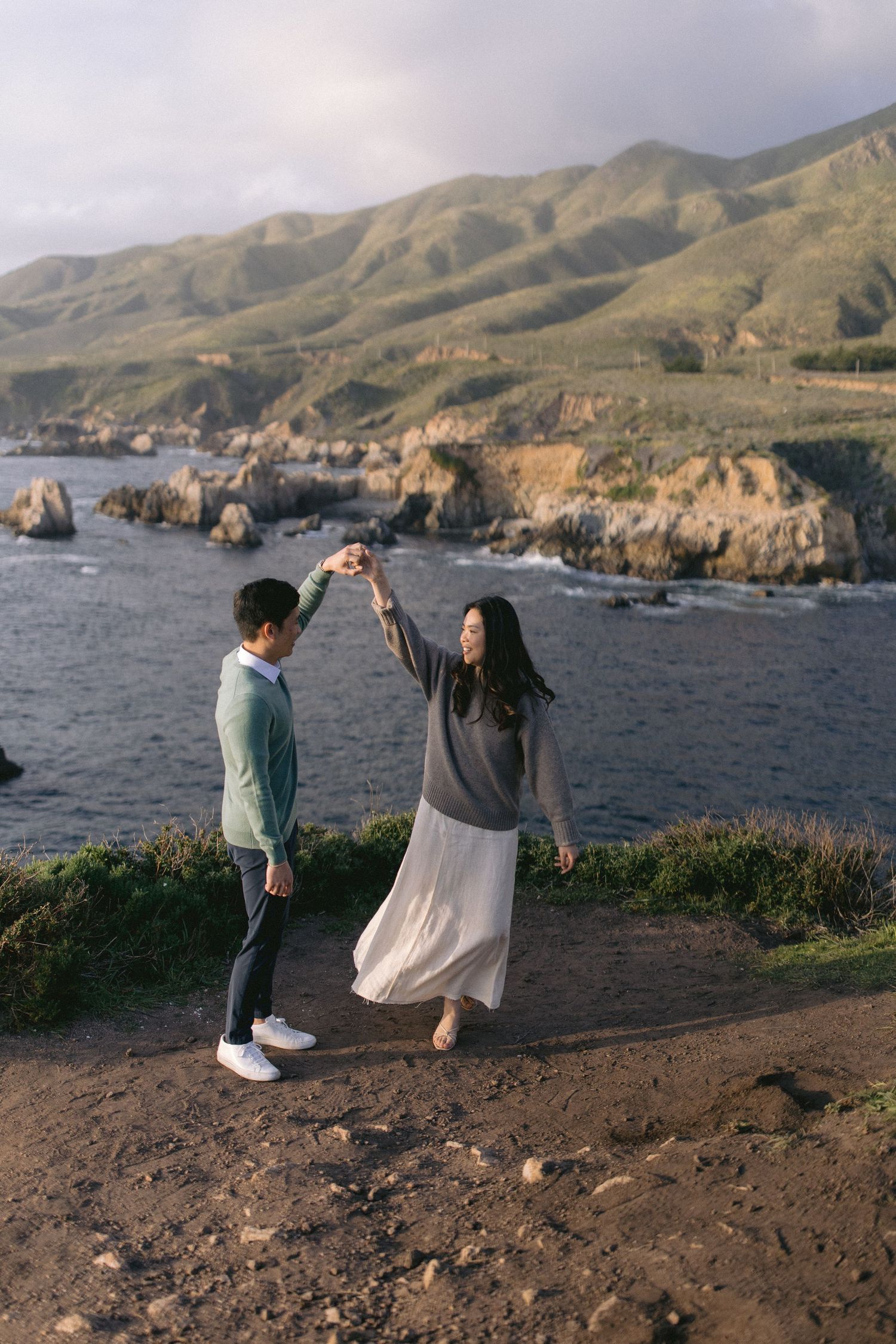 Couple dancing atop coastal cliffs overlooking rugged Big Sur coastline with mountains and rocky shores at sunset.