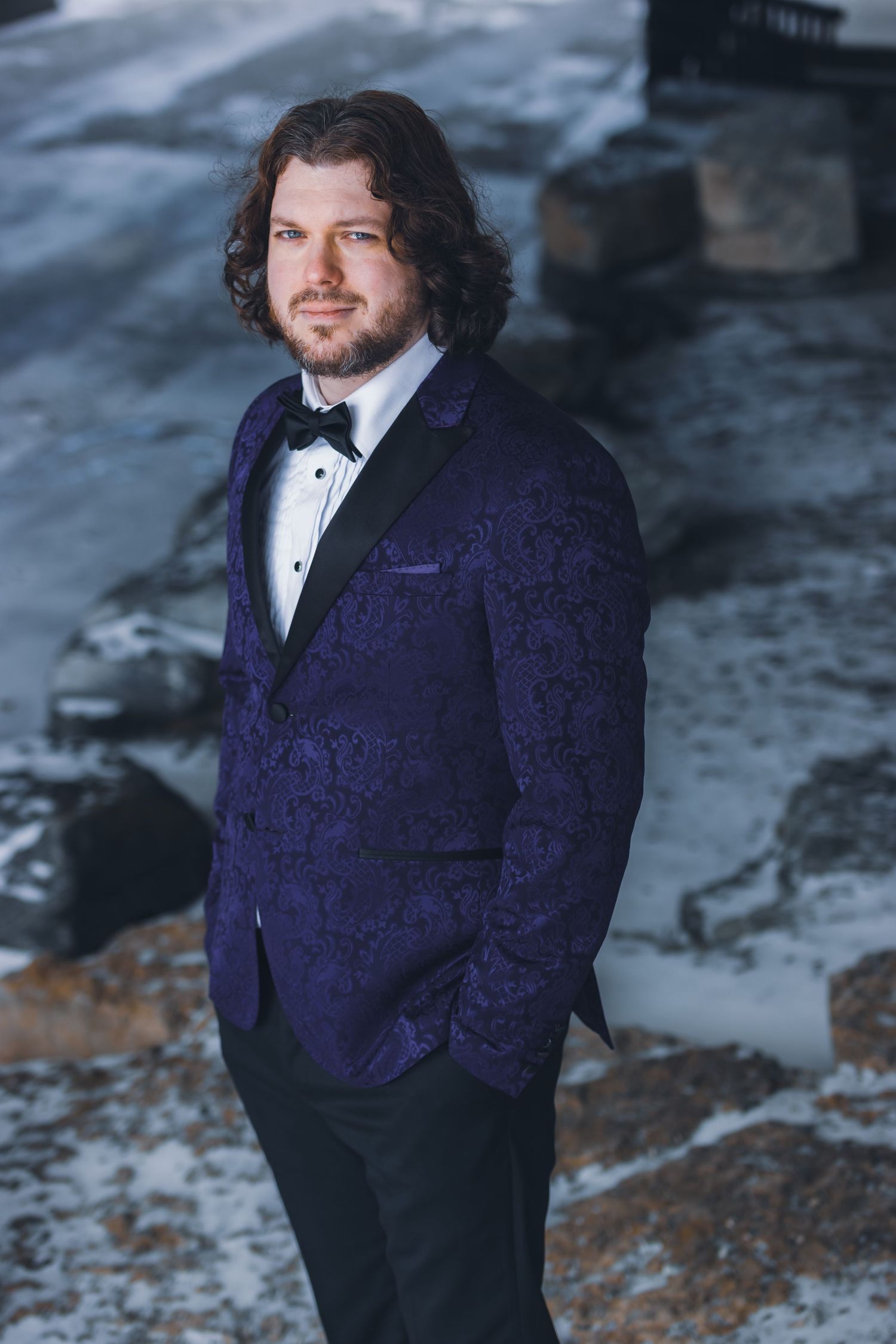 A formal portrait of someone wearing a blue velvet tuxedo with a bowtie standing against a snowy background.