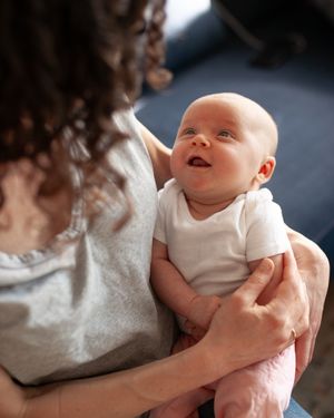 A smiling baby wearing a white onesie lies cradled in someone's arms looking up with joyful expression.