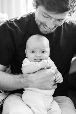 Black and white photo of a baby in white onesie being held and looking at camera with a happy expression while adult smiles.