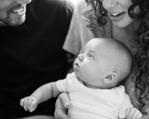 A tender black and white portrait of a tiny baby nestled between two adults, gazing up with curiosity.