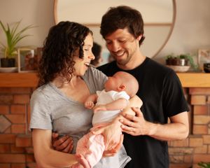 Two parents share a tender moment while holding their newborn baby in front of a brick fireplace at home.