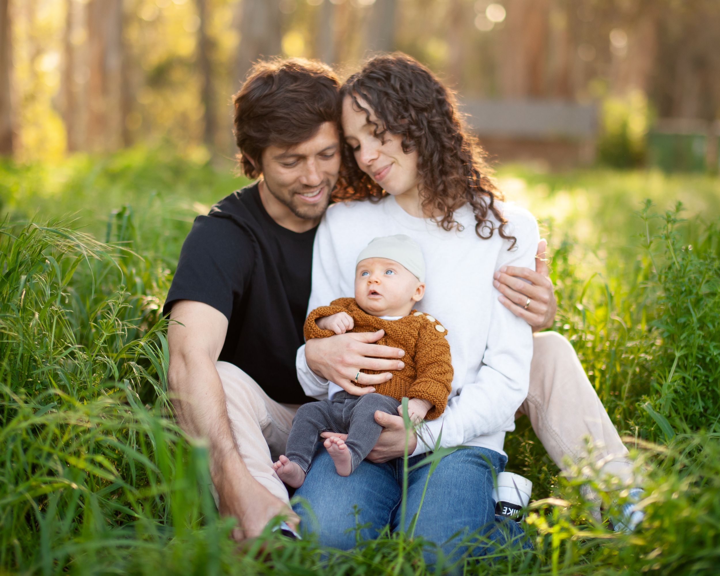 A couple sits in green grass at sunset with their baby wearing a brown top during an outdoor family photoshoot.