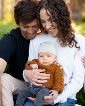 Happy family with a small baby wearing a brown knit sweater and white hat sitting together outdoors on a sunny autumn day.