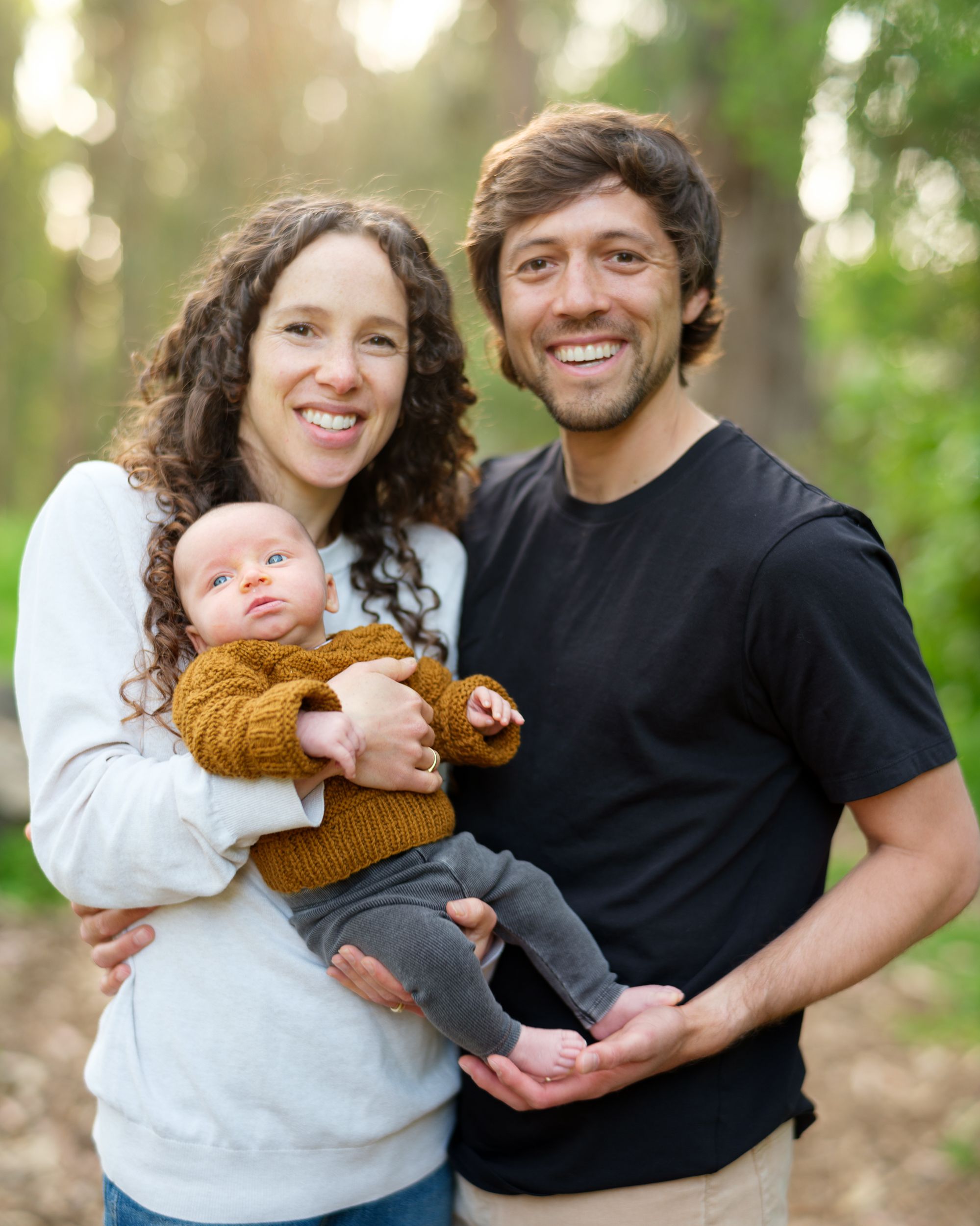 Family outdoors in nature holding baby wearing mustard sweater on sunny day with bokeh background.