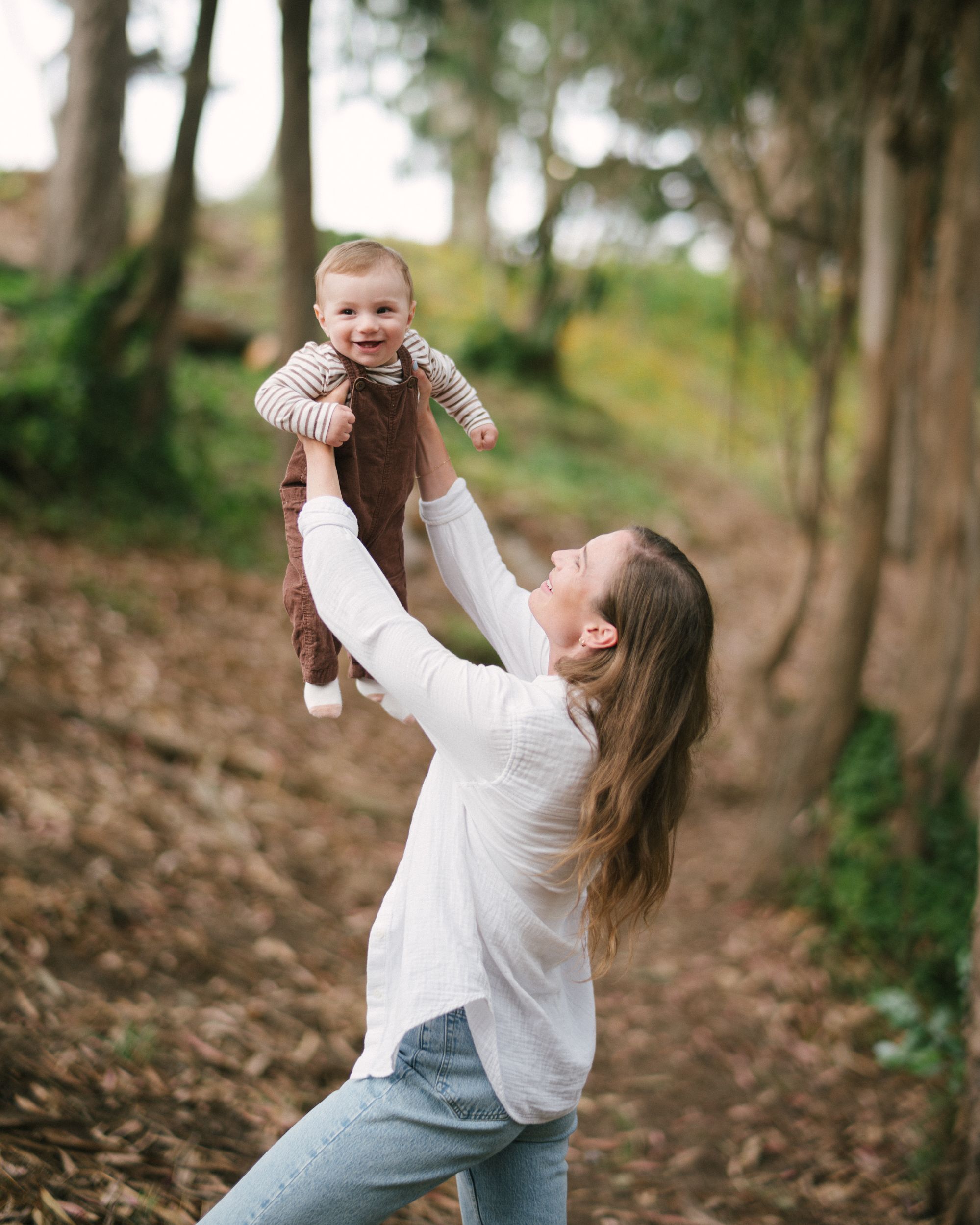 A parent lifts a small child wearing a brown outfit into the air while standing among trees in a wooded area.