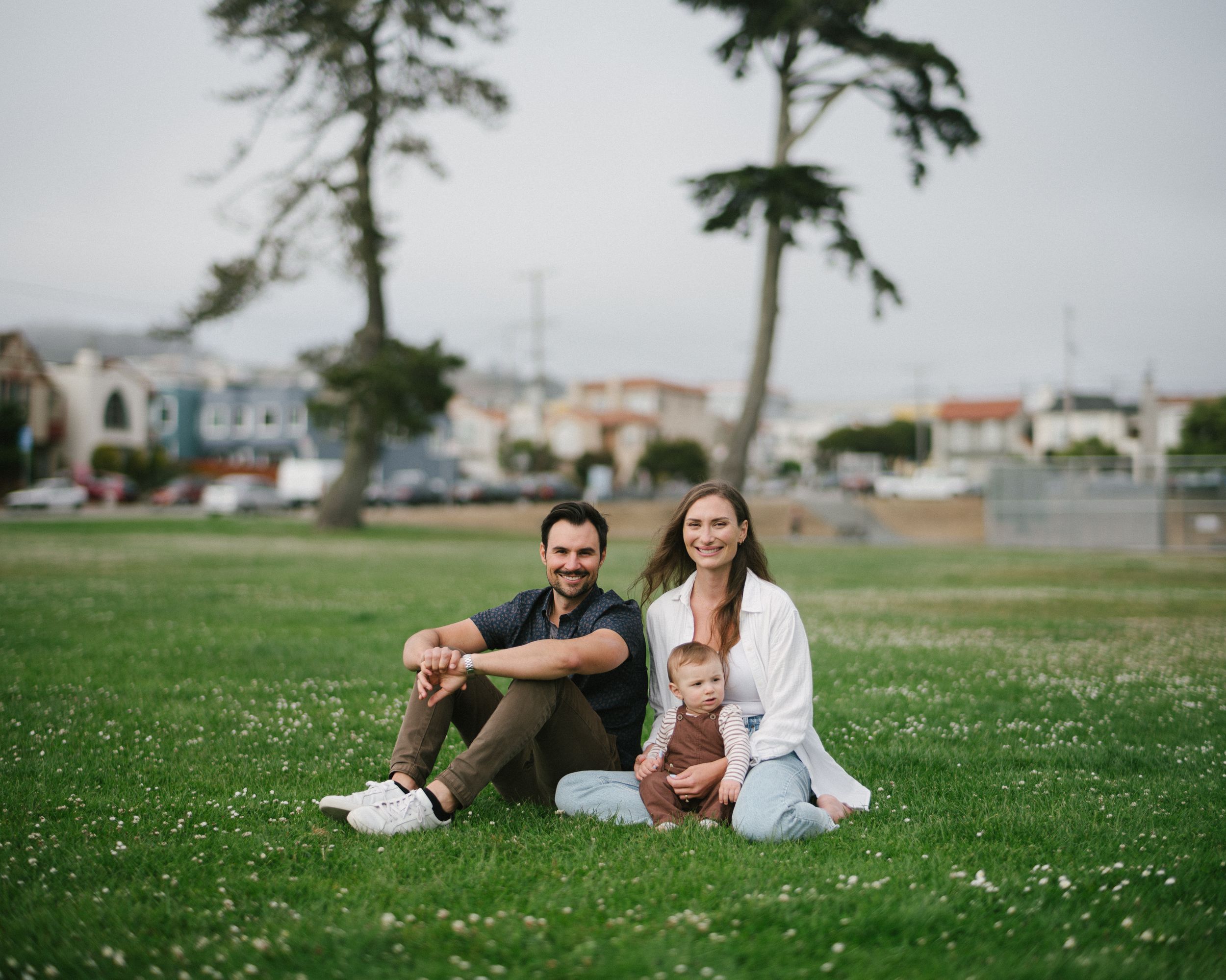 Family sitting together on green grass with coastal homes and tall pine trees in the background.