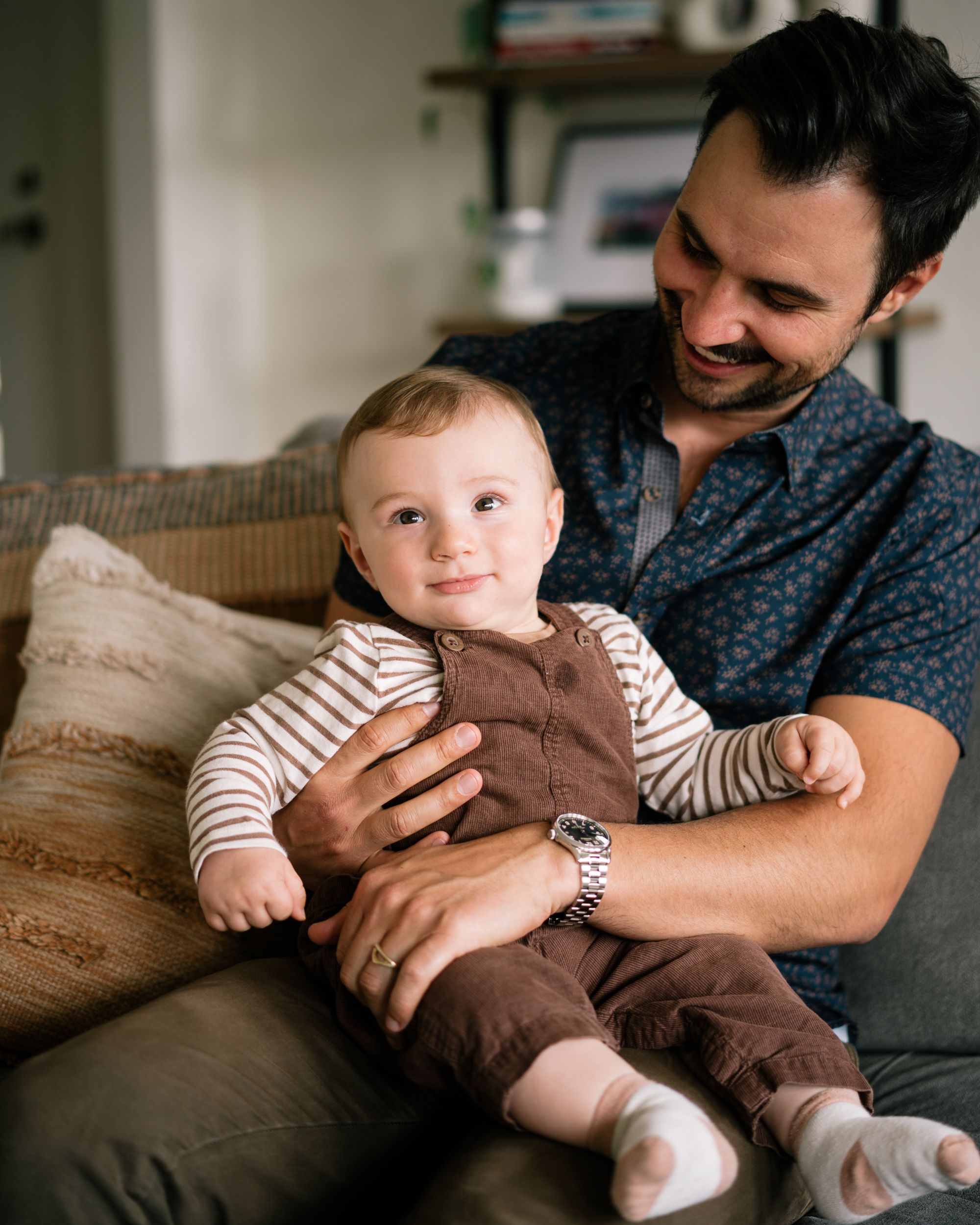 Candid family moment showing playful interaction with baby in brown overalls on grey couch.