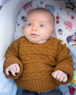 Baby wearing a cozy knit brown sweater lies on patterned bedding with bright blue eyes and sweet expression.