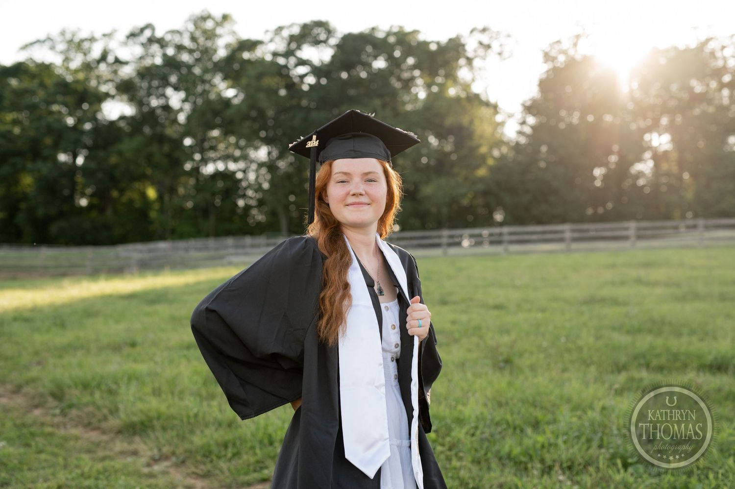 Graduate in cap and gown enjoys sunset moment in pastoral setting.