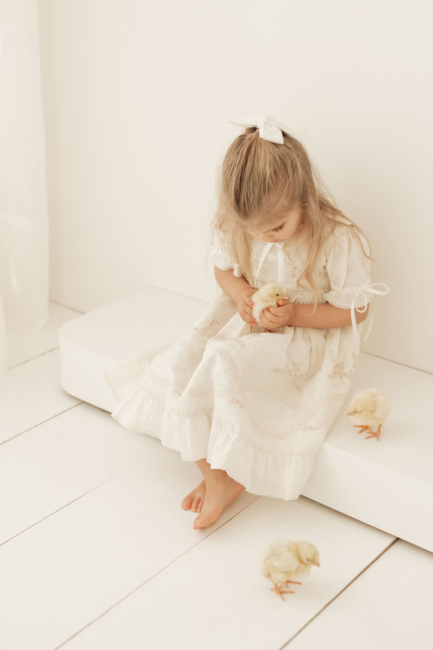 Child in white vintage dress sits on light floor with small duckling in soft, ethereal setting.