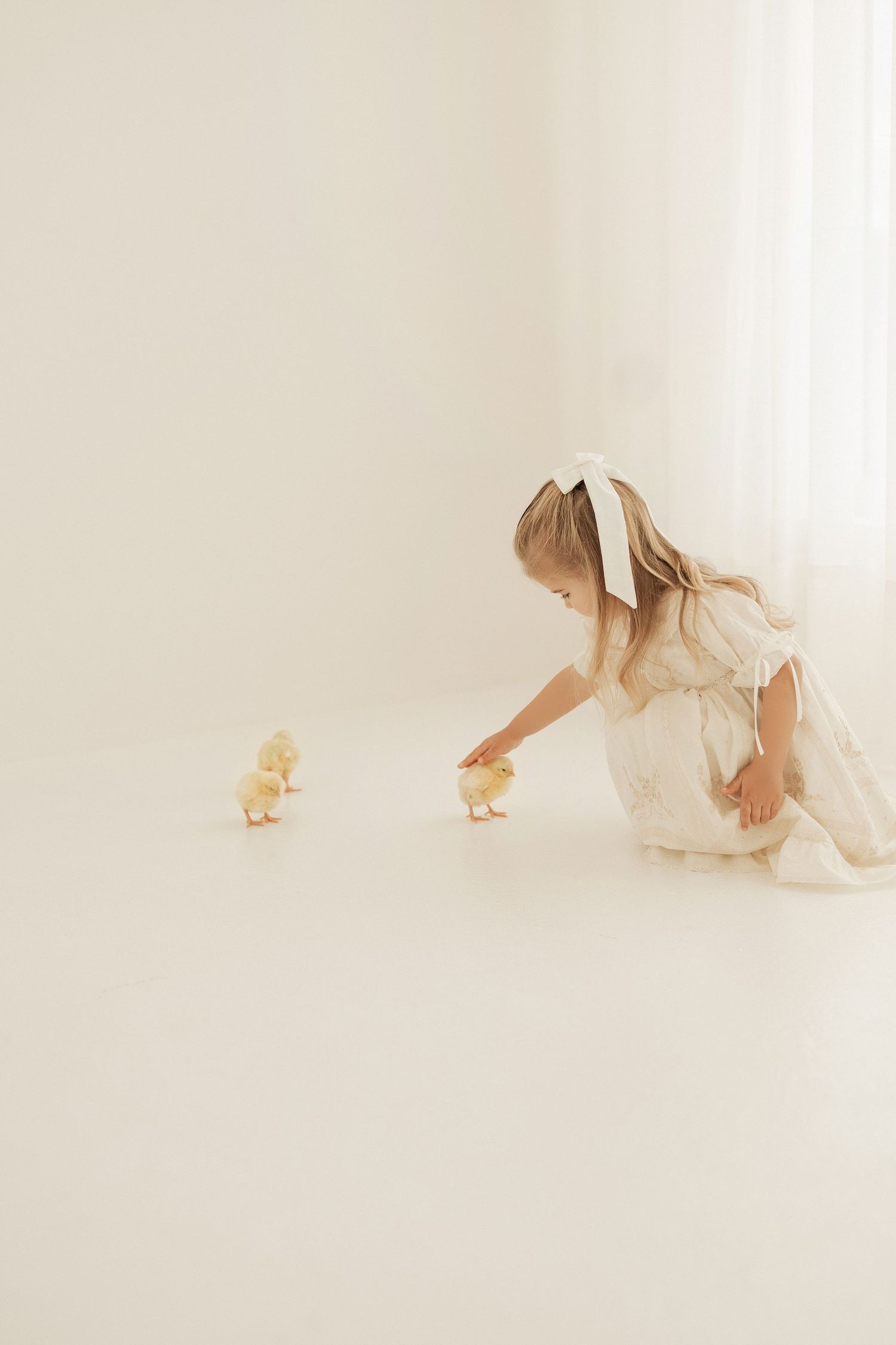 A person in a white dress reaches down to interact with two baby chicks in a bright minimalist setting.