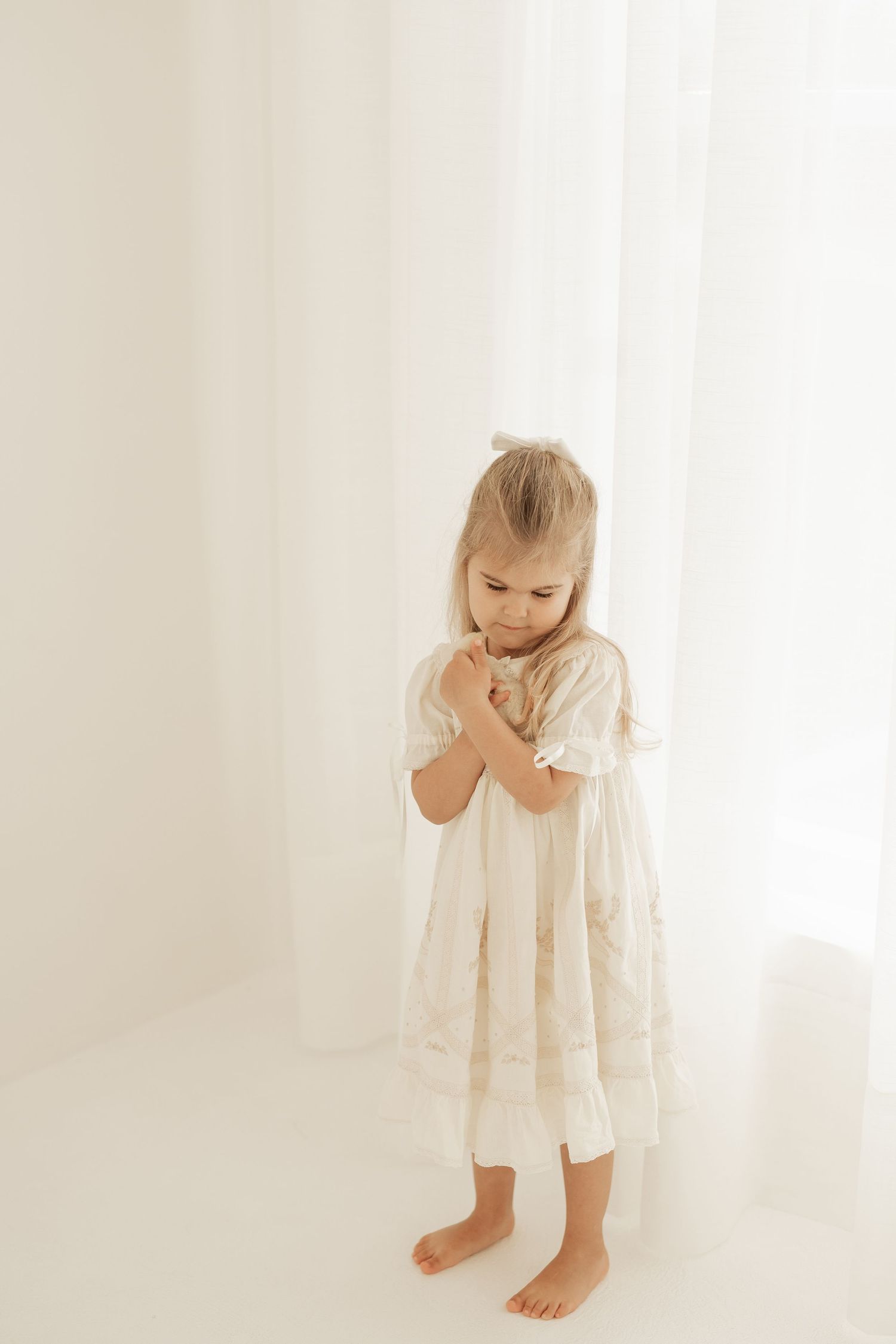 A young child stands barefoot wearing a white flowing nightgown against a light background.