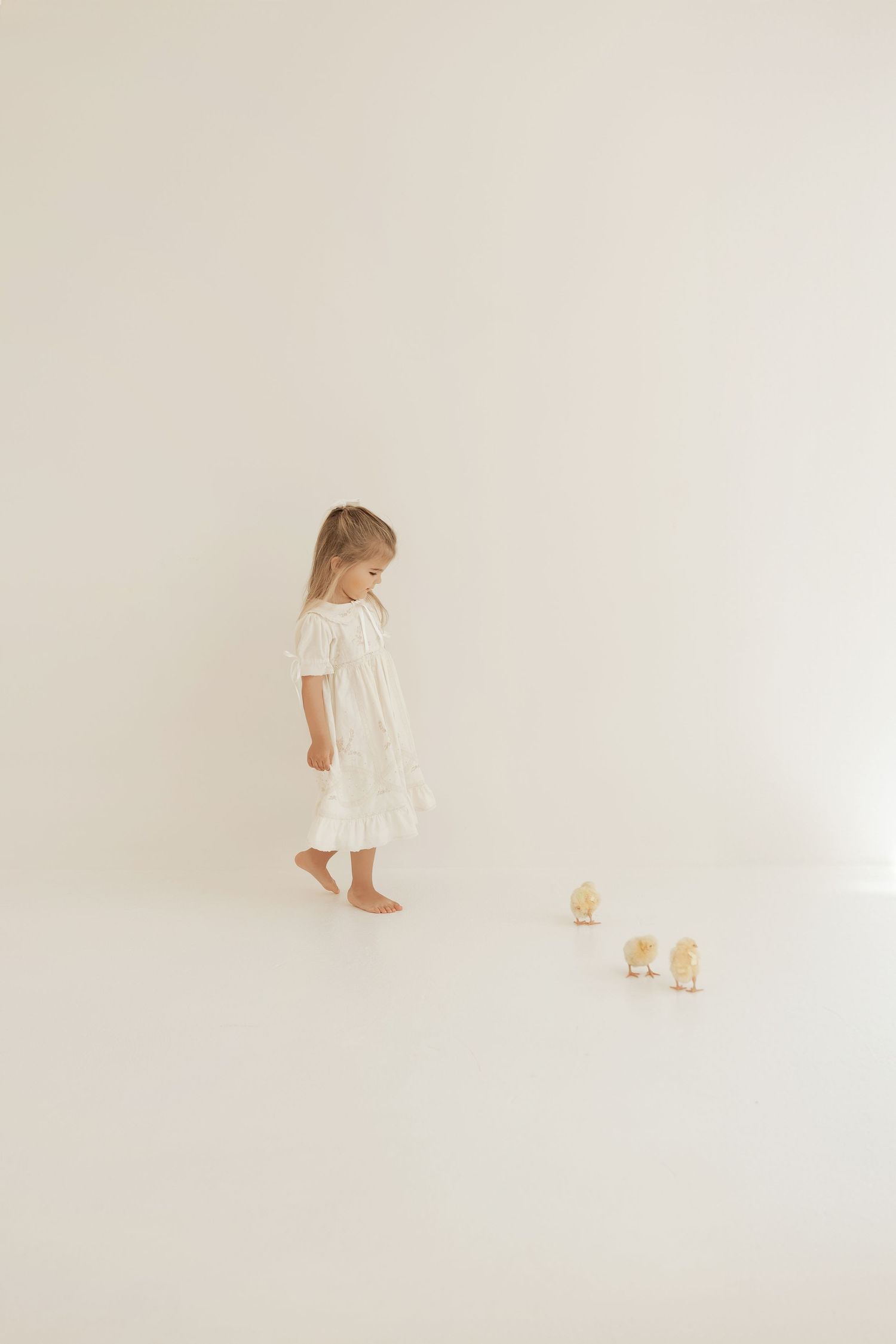 Child in white dress follows small yellow toy ducks across minimalist white background.