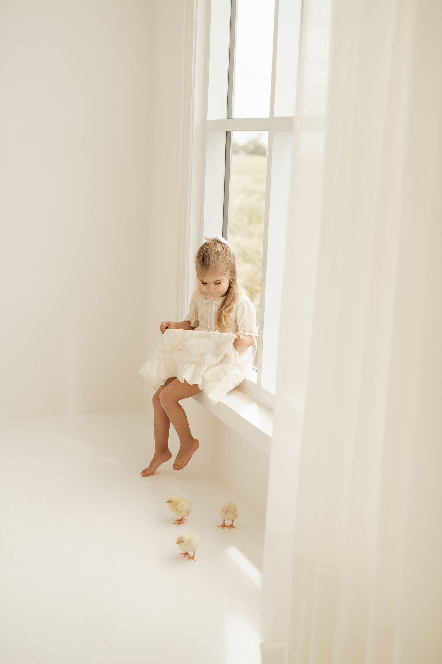 Child in white dress sits on bright windowsill with sunlight streaming in as baby chicks play below.