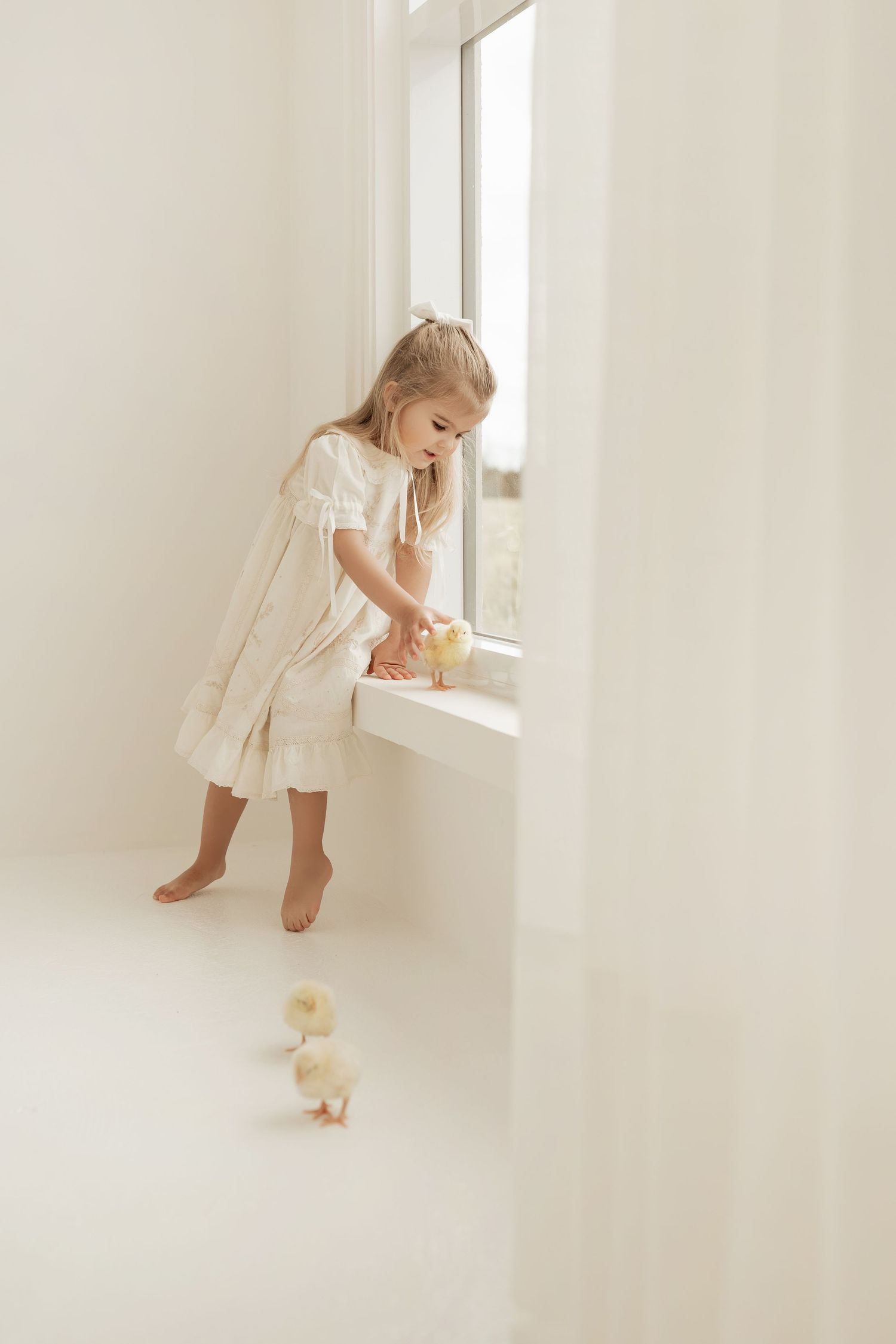 Child in a white nightgown stands by a bright window with a small toy duck on the floor.