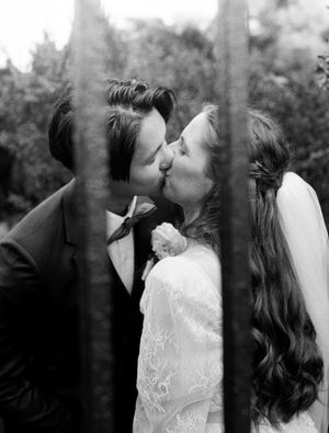A bride and groom share a tender kiss in a black and white vintage film photo framed by tree branches outdoors.