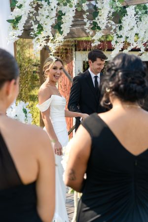 Bride laughing joyfully at outdoor wedding ceremony under white floral arch with groom in black suit.