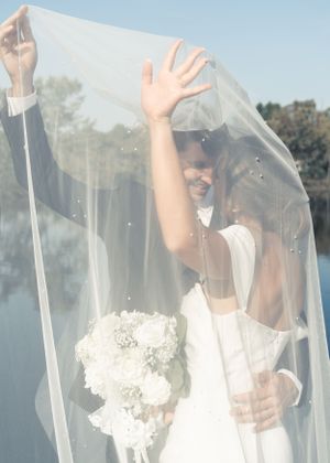 Bride and groom sharing romantic kiss under flowing white veil outdoors on wedding day.