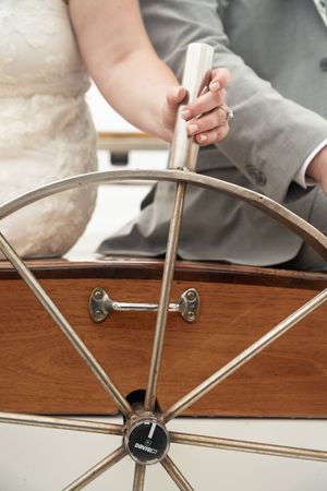 A bride and groom hold a sailboat steering wheel together, showcasing wedding rings during their nautical microwedding.
