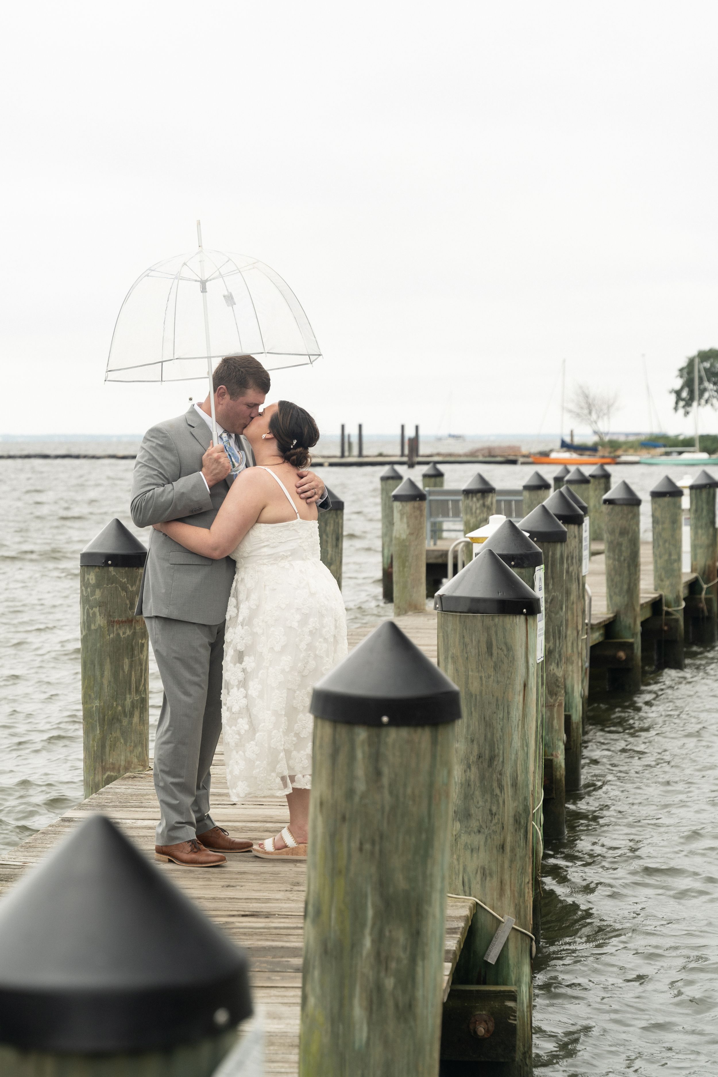 A bride and groom share a romantic kiss under a clear umbrella on a wooden dock by the water.