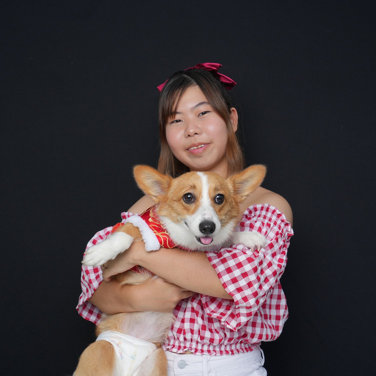 Person in red and white checkered top hugs a happy Pembroke Welsh Corgi against dark background.